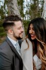 Side view of cheerful young bearded groom in suit laughing and cuddling elegant ethnic bride in white dress while standing together on wooden bridge near aged house in autumn forest — Stock Photo