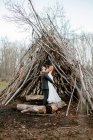 Side view of unrecognizable young groom hugging graceful bride in white wedding dress while standing in forest near twigs hut on cloudy day — Stock Photo