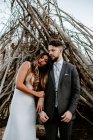 Молодий наречений обіймає витончену наречену в білій весільній сукні, стоячи в лісі біля гілки хатини в хмарний день — стокове фото