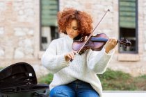 Giovane bella musicista donna in abbigliamento casual suonare il violino e guardando la fotocamera con calma mentre seduto su strada asfaltata — Foto stock