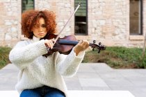 Jovem músico bonito do sexo feminino em uso casual tocando violino e olhando para a câmera calmamente enquanto sentado na rua pavimentada — Fotografia de Stock