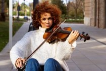 Jeune jolie musicienne en tenue décontractée jouant du violon et regardant calmement la caméra assise sur une rue pavée — Photo de stock