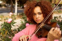 Músico feminino talentoso positivo com cabelo encaracolado vermelho usando suéter rosa tocando violino com os olhos fechados no parque ensolarado da cidade — Fotografia de Stock