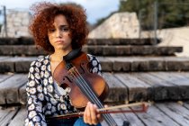 Junge hübsche Musikerin in Freizeitkleidung sitzt mit Geige auf der Uferpromenade und blickt gelassen in die Kamera im Sommerpark — Stockfoto