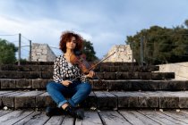 Full body young pretty female musician in casual wear sitting with violin on boardwalk and looking at camera calmly in summer park — Stock Photo