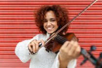 Feliz bonito músico feminino profissional em camisola branca tocando violino acústico e olhando para a câmera com sorriso de dente contra a parede vermelha — Fotografia de Stock