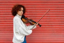 Felice bella musicista professionista femminile in maglione bianco suonare violino acustico e guardando la fotocamera con sorriso dentato contro la parete rossa — Foto stock