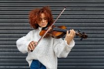 Jeune musicienne professionnelle poignante aux cheveux bouclés rouges portant un pull décontracté debout avec violon acoustique contre le mur du bâtiment et regardant la caméra — Photo de stock