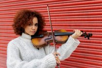 Feliz hermosa mujer profesional músico en suéter blanco tocando violín acústico y mirando a la cámara con sonrisa dentada contra la pared roja - foto de stock