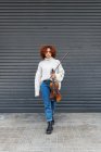 Jovem músico feminino profissional de comprimento completo com cabelo encaracolado vermelho vestindo camisola casual de pé com violino acústico contra a parede do edifício e olhando para a câmera — Fotografia de Stock