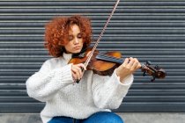 Wehmütige junge Profimusikerin mit rot gelockten Haaren in lässigem Pullover, die mit akustischer Geige vor einer Hauswand steht und in die Kamera blickt — Stockfoto