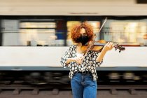 Привлекательная спокойная женщина-музыкант в повседневной одежде и защитной маске для лица, играющая на скрипке и смотрящая в камеру, стоя на платформе вокзала против отходящего поезда — стоковое фото