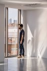 Seitenansicht eines Mannes, der am Fenster steht, in einem leeren, geräumigen Büroflur mit Schatten und Sonnenlicht an weißen Wänden und wegschaut — Stockfoto