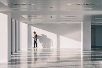Вид сбоку мужчины, стоящего в пустом офисном коридоре с белыми стенами и творческими тенями при использовании мобильного телефона — стоковое фото