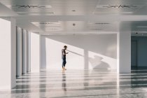 Vista lateral do macho em pé no corredor do escritório vazio com paredes brancas e sombras criativas ao usar o telefone móvel — Fotografia de Stock