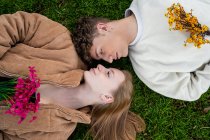 Visão aérea do jovem casal olhando um para o outro enquanto descansa na grama com buquês de flores florescendo — Fotografia de Stock