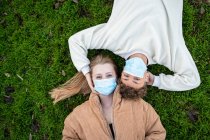 Visão superior do jovem casal anônimo em máscaras respiratórias deitado no prado enquanto olha para a câmera durante a pandemia de COVID 19 — Fotografia de Stock