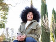 Niedriger Winkel der charismatischen jungen afroamerikanischen Millennials mit lockigem Haar, die lachen und wegschauen, während sie an sonnigen Tagen ihr Smartphone im Park benutzen — Stockfoto