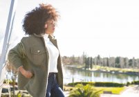 Fröhliche junge Afroamerikanerin mit lockigem Haar in stylischem warmem Outfit lächelt und blickt in die Kamera, während sie sich an einem sonnigen Herbsttag im Park am Seeufer ausruhen — Stockfoto