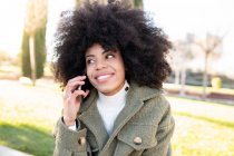 Low angle of charismatic young African American female millennial with curly hair laughing and looking away while using smartphone in park on sunny day — Stock Photo