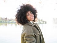 Joven y alegre hembra afroamericana con el pelo rizado en un elegante atuendo cálido sonriendo y mirando a la cámara mientras descansa en el parque junto al lago en el soleado día de otoño - foto de stock