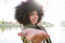 Seitenansicht einer positiven jungen schwarzen Dame mit Afro-Frisur im trendigen Outfit, die die Hand in Richtung Kamera ausstreckt und lächelt, während sie sich in der Nähe des Sees im Herbstpark ausruht — Stockfoto