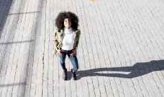 Von oben eine ausdrucksstarke junge schwarze Dame mit Afro-Frisur in stylischer Kleidung, die an sonnigen Tagen auf gepflasterten Straßen steht und lächelt — Stockfoto