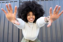 Joyeux jeune afro-américaine dame avec les cheveux bouclés dans des vêtements décontractés montrant deux mains et regardant la caméra sur la rue — Photo de stock