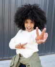 Веселая молодая афроамериканка с кудрявыми волосами в повседневной одежде показывает два пальца знак с открытым ртом и смотрит в камеру на улице — стоковое фото