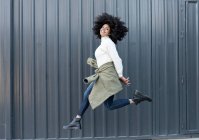 Vista lateral da jovem mulher afro-americana feliz com cabelo encaracolado na roupa da moda sorrindo e olhando para a câmera enquanto pulava na rua — Fotografia de Stock