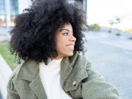 Trendy young black female millennial with Afro hair in stylish warm clothes resting on street and looking away pensively near modern building with glass walls — Stock Photo