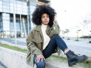 Moda joven negro femenino millennial con pelo afro en ropa de abrigo elegante descansando en la calle y mirando hacia otro lado pensativamente cerca de un edificio moderno con paredes de vidrio - foto de stock