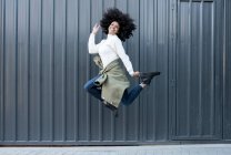 Vista laterale della giovane donna afroamericana felice con i capelli ricci in abito alla moda sorridente e guardando la fotocamera mentre salta sulla strada — Foto stock