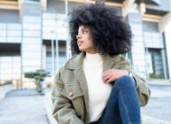 Moda jovem preto feminino millennial com cabelo afro em roupas quentes elegantes descansando na rua e olhando para longe pensivamente perto de edifício moderno com paredes de vidro — Fotografia de Stock