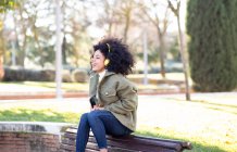 Aufgeregte junge Afroamerikanerin mit lockigem Haar in stylischem Outfit, die auf Bank im Park ruht und Lieblingslied über Kopfhörer hört — Stockfoto