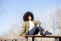 Cuerpo completo de mujer afroamericana joven reflexiva con cabello rizado oscuro en ropa de abrigo de moda relajándose en el banco de madera en el parque y mirando hacia otro lado soñando en el soleado día de otoño - foto de stock