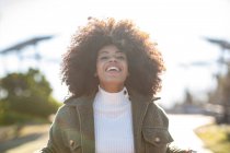 Joven y alegre hembra afroamericana con el pelo rizado en un elegante atuendo cálido sonriendo y mirando a la cámara mientras descansa en el parque junto al lago en el soleado día de otoño - foto de stock