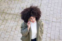 Високий кут зосередженої молодої афроамериканської жінки тисячоліття з кучерявим волоссям в стильному одязі, який розмовляє по телефону під час ходьби по брукованій вулиці в сонячний день. — стокове фото