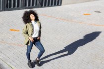 Von oben eine ausdrucksstarke junge schwarze Dame mit Afro-Frisur in stylischer Kleidung, die an sonnigen Tagen auf gepflasterten Straßen steht und lächelt — Stockfoto