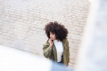 Высокий угол концентрированной молодой афроамериканской девушки тысячелетия с кудрявыми волосами в стильном наряде, разговаривающей по телефону во время прогулки по асфальтированной улице в солнечный день — стоковое фото