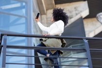 Seitenansicht einer selbstbewussten jungen schwarzen Frau mit Afro-Frisur in lässiger Kleidung, die lächelt, während sie ein Selfie auf dem Smartphone macht, das auf dem Balkon eines modernen Gebäudes steht — Stockfoto