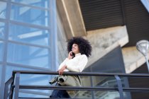 Vista lateral da jovem mulher negra confiante com penteado afro em roupas casuais sorrindo enquanto conversa no smartphone em pé na varanda do edifício moderno — Fotografia de Stock