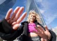 Снизу веселая женщина с кудрявыми светлыми волосами и в причудливом наряде протягивает руки к камере, стоя в центре города против небоскребов в солнечный день — стоковое фото