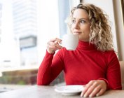 Dreamy female sitting at table in cafe and drinking hot beverage while enjoying weekend and looking out of window — Stock Photo