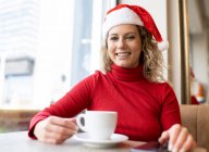 Mujer encantada en Santa sombrero y cuello alto rojo sentado con taza de café en la cafetería y mirando a la cámara durante las vacaciones de Navidad - foto de stock