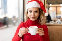 Приємна жінка в Санта-Каті і червона білизна сидять з чашкою кави в кафе і дивляться на камеру під час різдвяних свят. — стокове фото