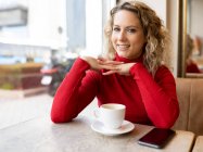 Мечтательная женщина сидит за столом в кафе и пьет горячий напиток, наслаждаясь выходными и глядя в камеру — стоковое фото
