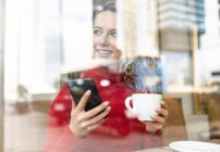 Через вікно весела жінка зі смартфоном і чашкою гарячого напою, сидячи за столом в кафе і дивлячись геть — стокове фото