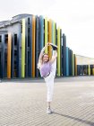 Full length attractive fit female in casual wear performing standing split and looking at camera with smile while standing on against creative building on sunny summer day — Stock Photo