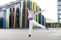 Full length happy young fit female wearing casual clothes jumping with arms raised against creative modern building and looking away with smile on clear summer weather — Stock Photo
